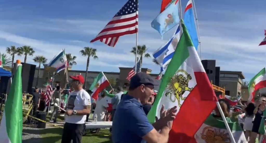 Crowd at an outdoor festival waving American and Mexican flags under a blue sky with palm trees in the background.