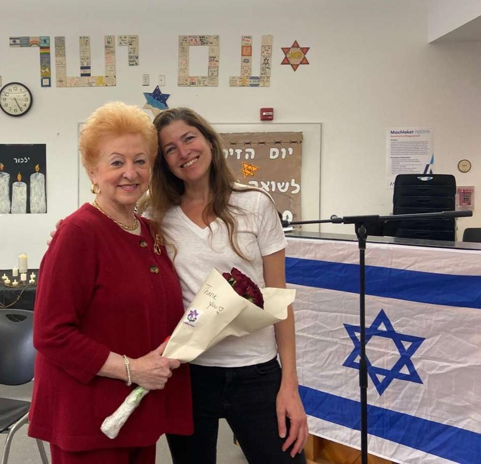Two women smiling and posing together; an older woman in red holds a bouquet while standing beside a younger woman in a white shirt in a room decorated with an Israeli flag and Star of David symbols.